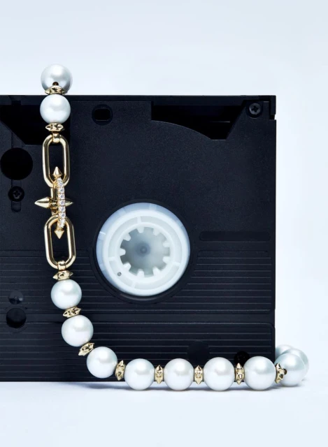 A pearl necklace with gold accents is draped over a black VHS tape against a white background in a studio setting, showcasing fashion jewelry.