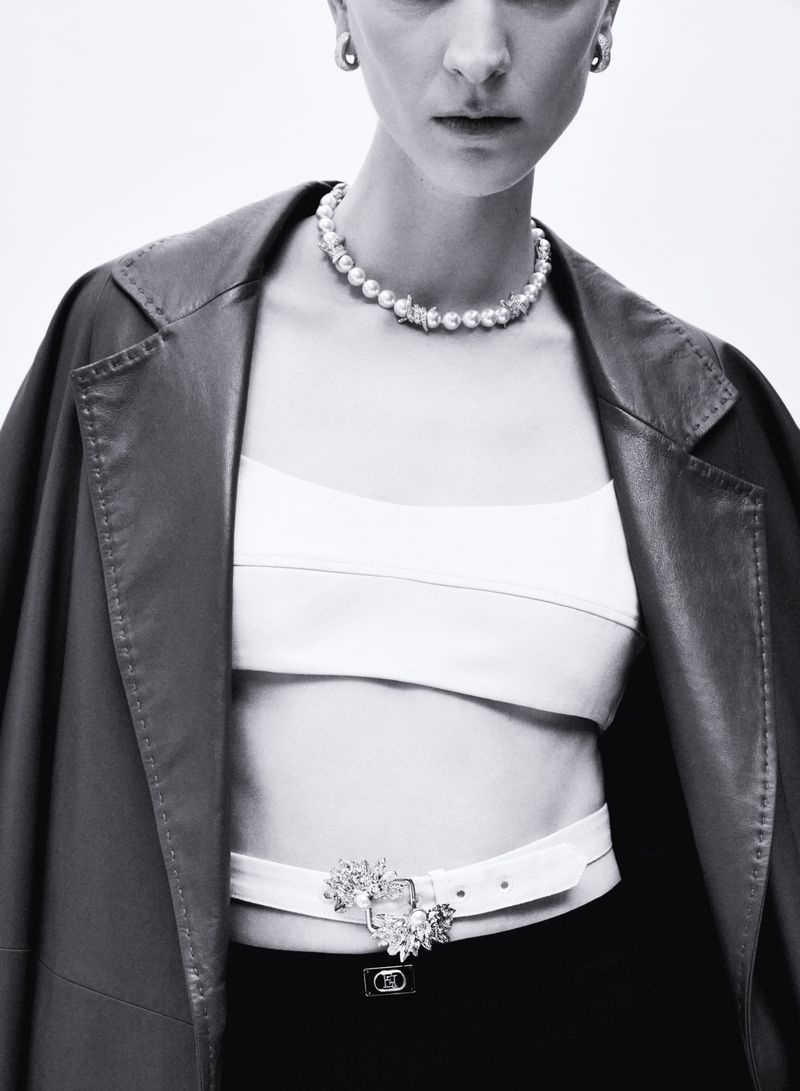 Model wearing dark leather jacket, white crop top, and pearl necklace against white background