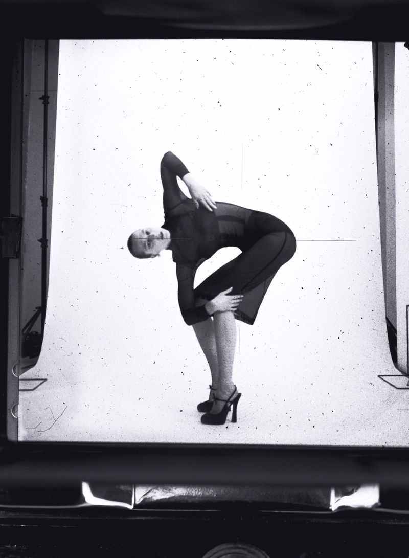 Black and white photograph of a woman in a dark outfit, angled backward in a dance-like pose against a bright, textured backdrop.