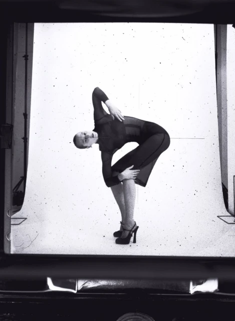 Black and white photograph of a woman in a dark outfit, angled backward in a dance-like pose against a bright, textured backdrop.