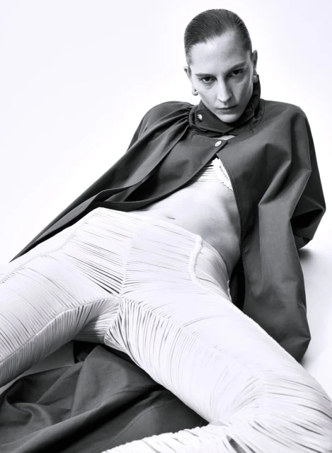 Fashion shot of a woman with slicked-back hair reclining and looking at the camera, wearing a dark jacket and light textured pants.