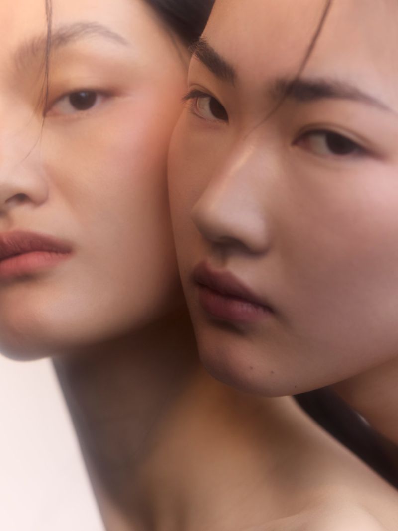 Close-up portrait showcasing the faces of two Asian adult females, set against a soft, blurred light backdrop, emphasizing their natural beauty.
