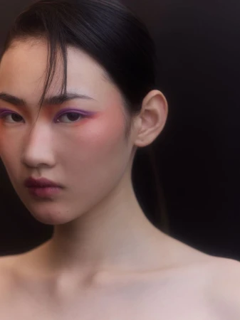 Close-up portrait of an East Asian woman with purple and orange eye makeup against a dark background.