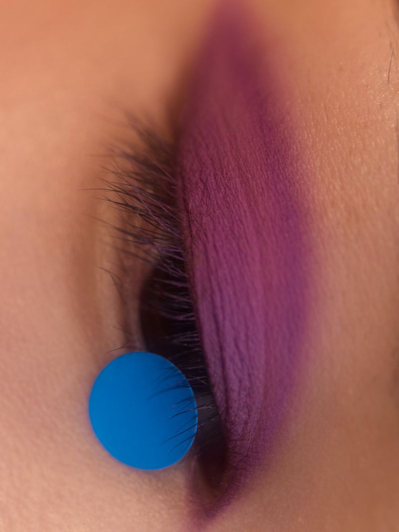 Close-up of an eye with purple eyeshadow and long lashes, a small blue dot accentuates the beauty look, creating a striking visual.