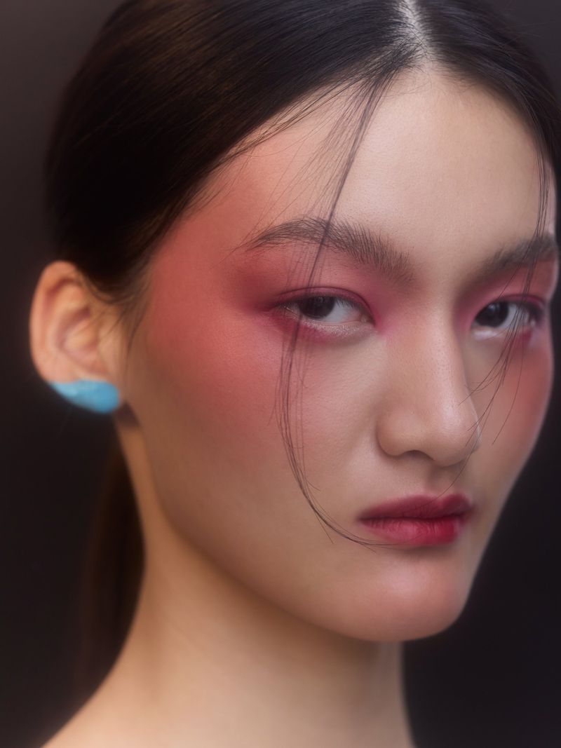 Headshot of an Asian female model showcasing vivid pink and red makeup on her face, lips, and eyebrows against a dark background.