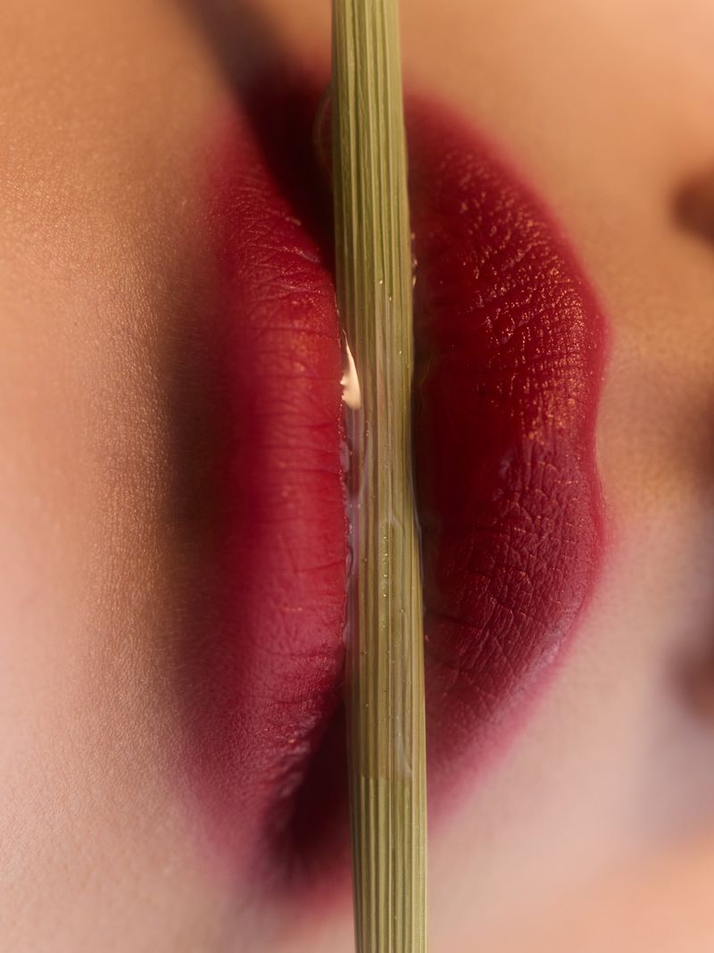 Close-up of a person's face with red lipstick, featuring lips and a vertical green reed or stem partially obscuring the face.