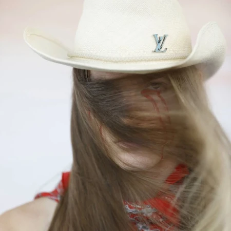 Woman with tears painted on face wearing a Louis Vuitton cowboy hat and red bandana against a neutral backdrop in a close-up studio shot.