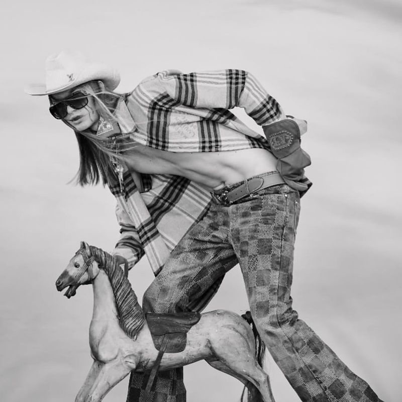 Fashion shot in black and white of person wearing cowboy hat, sunglasses, and flannel shirt while riding toy horse.