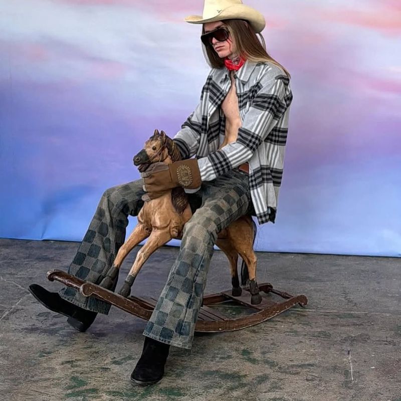 Adult male with a cowboy hat and sunglasses poses while riding a toy horse in a studio setting with a light purple background.