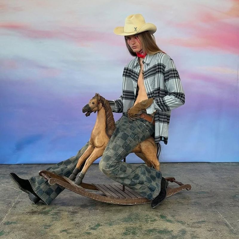 A person wearing a cowboy hat and a casual shirt sits on a wooden rocking horse with a sky-painted backdrop in a studio setting.