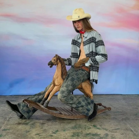 A person wearing a cowboy hat and a casual shirt sits on a wooden rocking horse with a sky-painted backdrop in a studio setting.