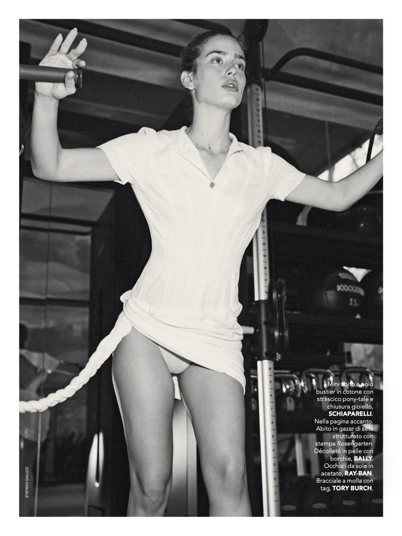 A slender young woman is standing indoors in a gym, holding onto a cable machine. She is wearing a short white dress in the black and white photograph.