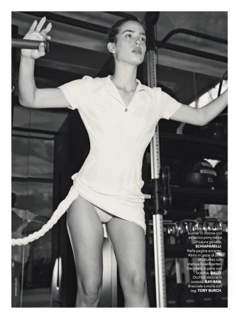 A slender young woman is standing indoors in a gym, holding onto a cable machine. She is wearing a short white dress in the black and white photograph.