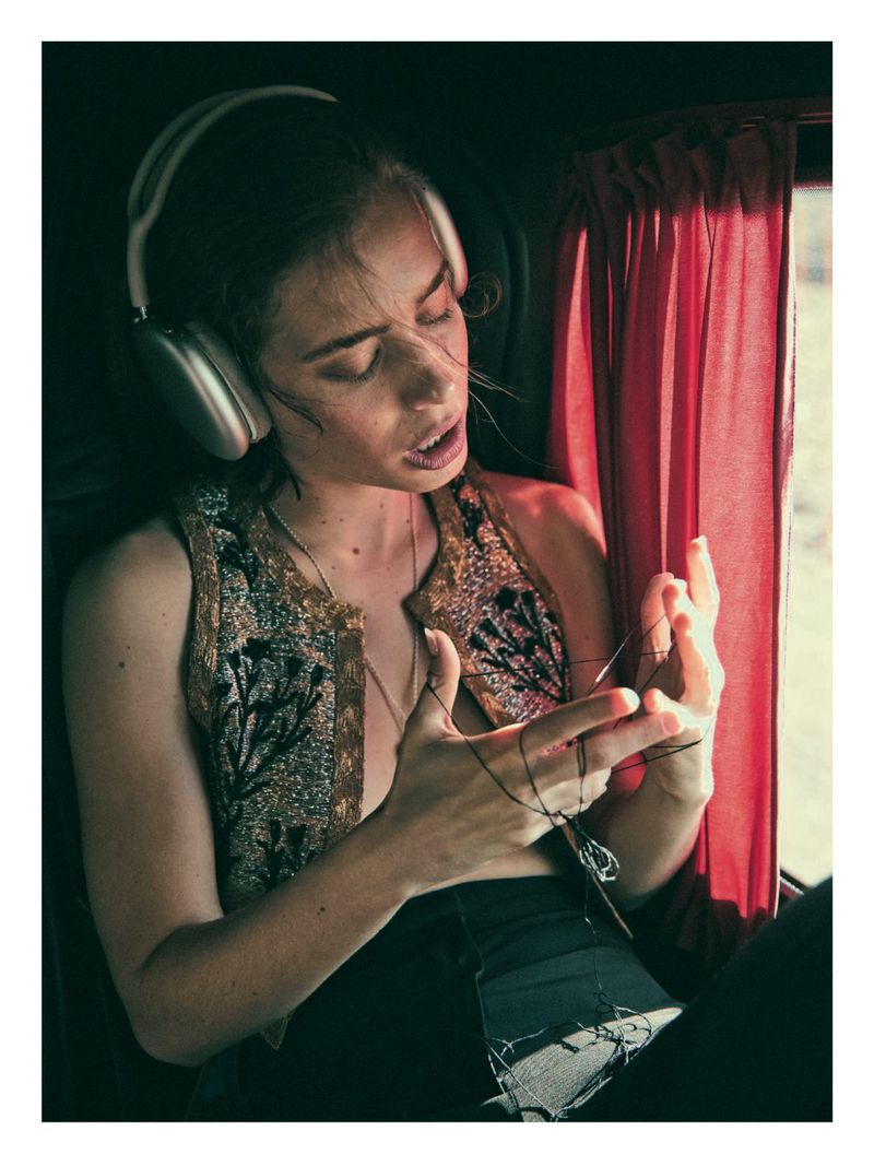 Portrait of a woman with headphones, wearing a decorated vest, holding a string indoors with window light and red curtain.