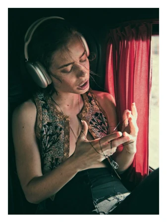 Portrait of a woman with headphones, wearing a decorated vest, holding a string indoors with window light and red curtain.