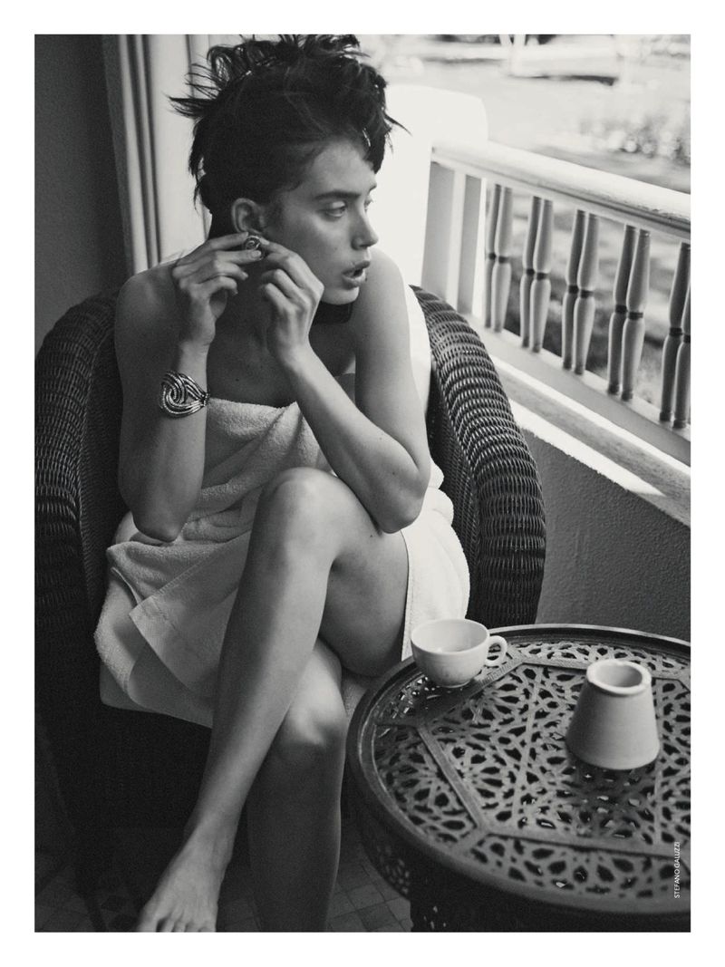 Model wrapped in white towel sitting on wicker chair adjusting earring on balcony in black and white