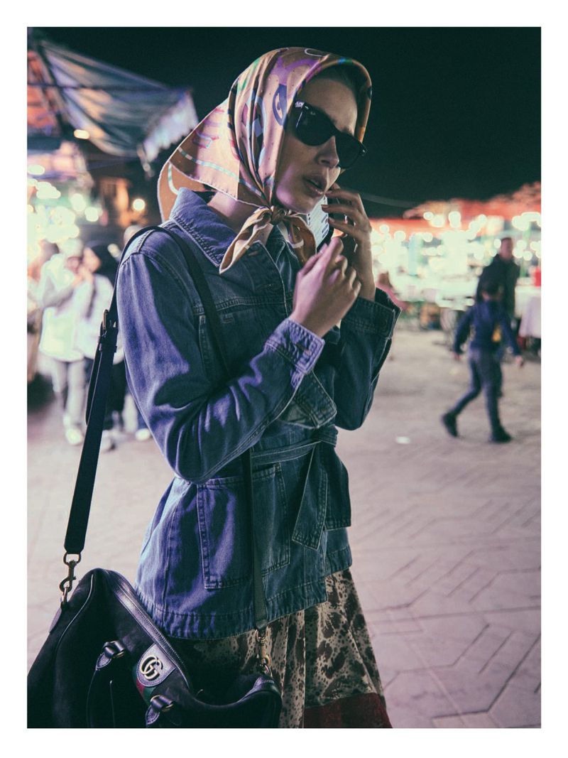 A woman in a headscarf, sunglasses, and denim jacket stands on a brick surface at night, holding a phone to her ear, with other people blurred in the background.