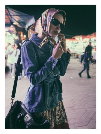 A woman in a headscarf, sunglasses, and denim jacket stands on a brick surface at night, holding a phone to her ear, with other people blurred in the background.