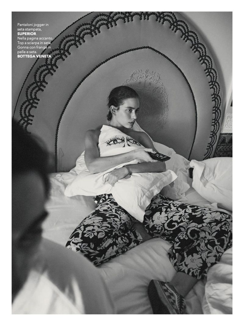 Black and white image of a woman in bed with a pillow, featuring a headboard, loungewear, and a person in the foreground.