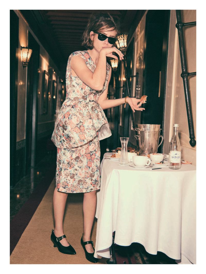 Woman in floral dress and sunglasses eats a snack at a table with refreshments in a hallway with sconces and dark paneling.