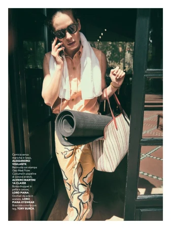 Adult male on the phone near an open door, with a towel, yoga mat, and tote bag, wearing sunglasses, a peach shirt, and patterned pants.