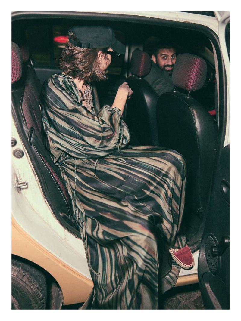 Woman in a patterned dress and baseball cap exits a car, with a man smiling in the back seat, creating a candid and urban atmosphere.