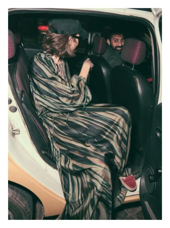 Woman in a patterned dress and baseball cap exits a car, with a man smiling in the back seat, creating a candid and urban atmosphere.