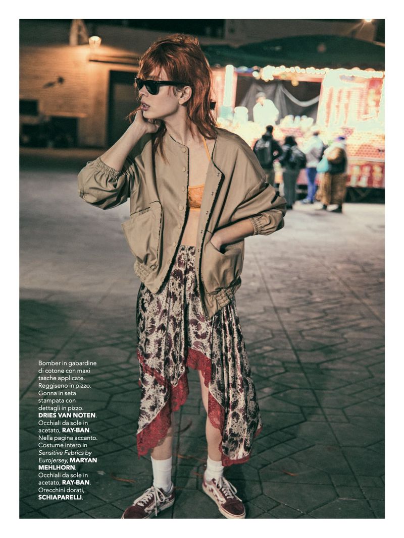 Fashion editorial shot of an adult woman in a bomber jacket, skirt, and sunglasses posing on a city street at night with blurred people in background.