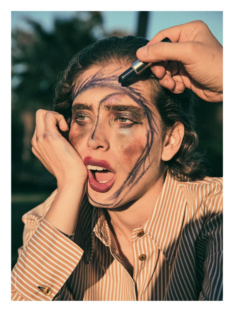 Woman with drawn-on makeup being touched up, wearing a striped shirt in a grassy outdoor setting with trees and warm lighting.