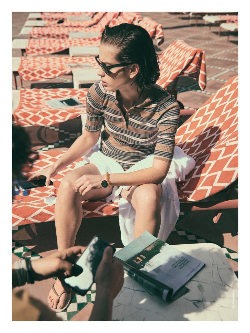 A woman wearing sunglasses and a striped top relaxing on a patterned lounge chair outdoors, with a magazine and phone on a table.