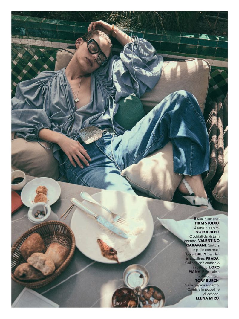 A fashion editorial shot of a woman reclining at a table with breakfast items, wearing a blouse, jeans, and sunglasses in a bright outdoor setting.