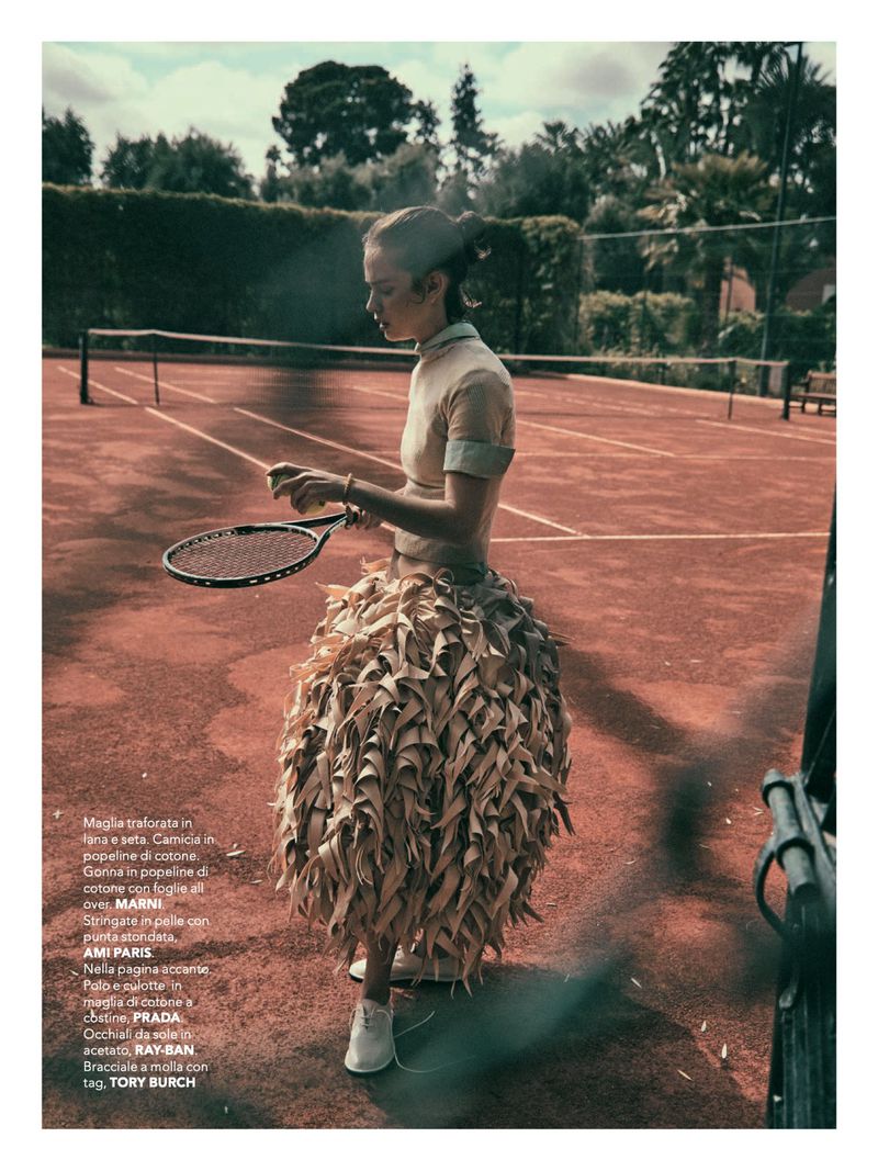 Woman in avant-garde fashion on a tennis court, holding a tennis ball and racket. Trees and a net visible in the background.