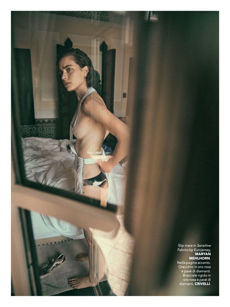 Woman poses in a bedroom wearing lingerie and a white robe. Reflected view through window with light coming through bedroom.