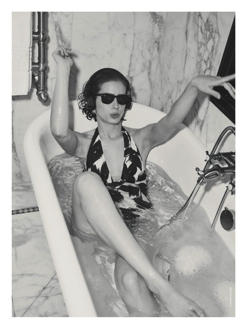 Woman in patterned swimsuit and sunglasses sits in bathtub with her arms raised in black and white photography; marble background.
