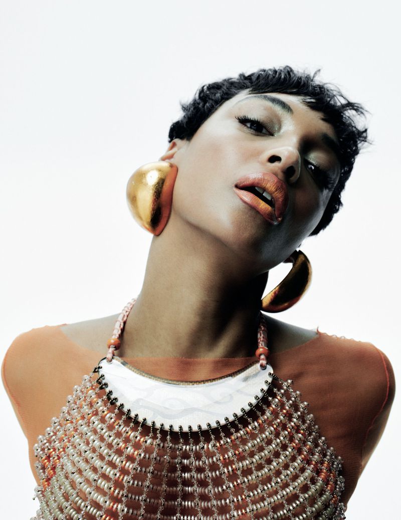 Headshot of an adult woman with short dark hair, large gold earrings, and beaded jewelry against a bright white background. Beauty portrait.