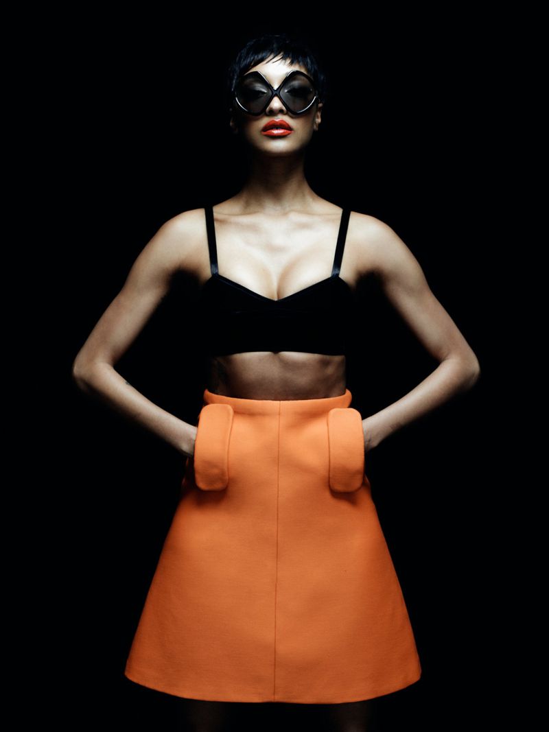 Jourdan Dunn wearing a black crop top and orange skirt with dark sunglasses against a black background