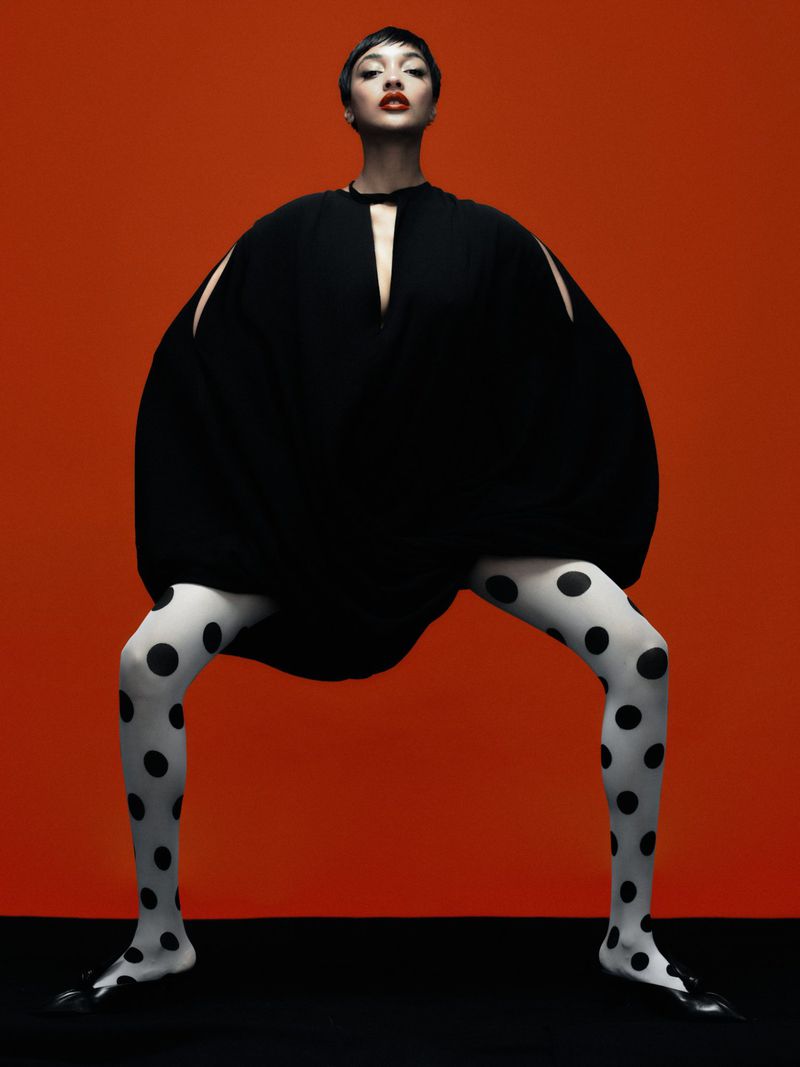 A fashion shot of a slender adult female model in a black dress with cutouts and patterned tights, posing on a black surface against a red backdrop.