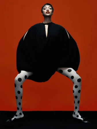 A fashion shot of a slender adult female model in a black dress with cutouts and patterned tights, posing on a black surface against a red backdrop.