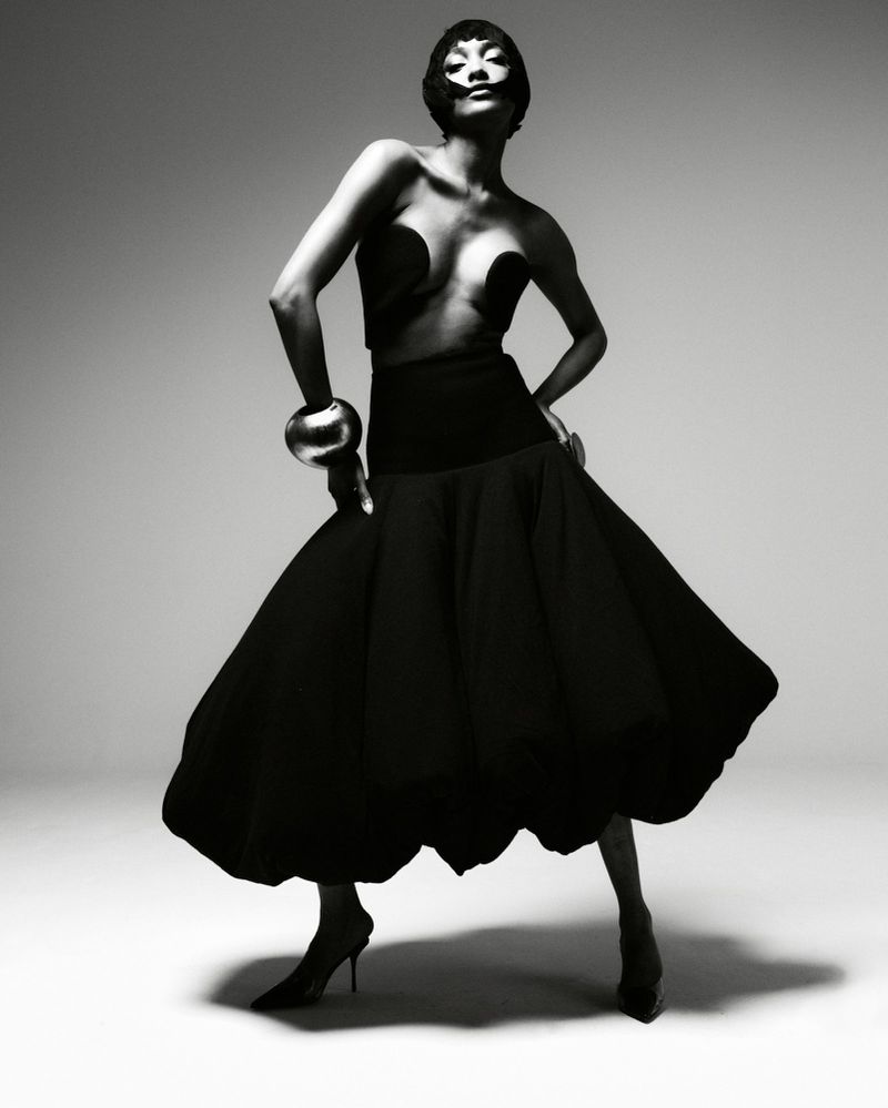 Full-length shot of a woman in black couture dress with sculptural details, accessorized with a wide silver bracelet, against gray studio backdrop.