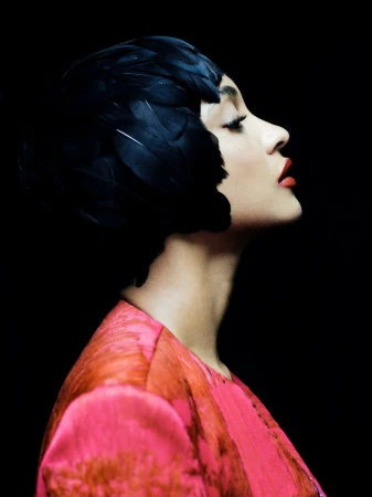 Profile portrait of a woman with red lipstick and a black feathered headpiece against a dark backdrop in a beauty or fashion editorial.