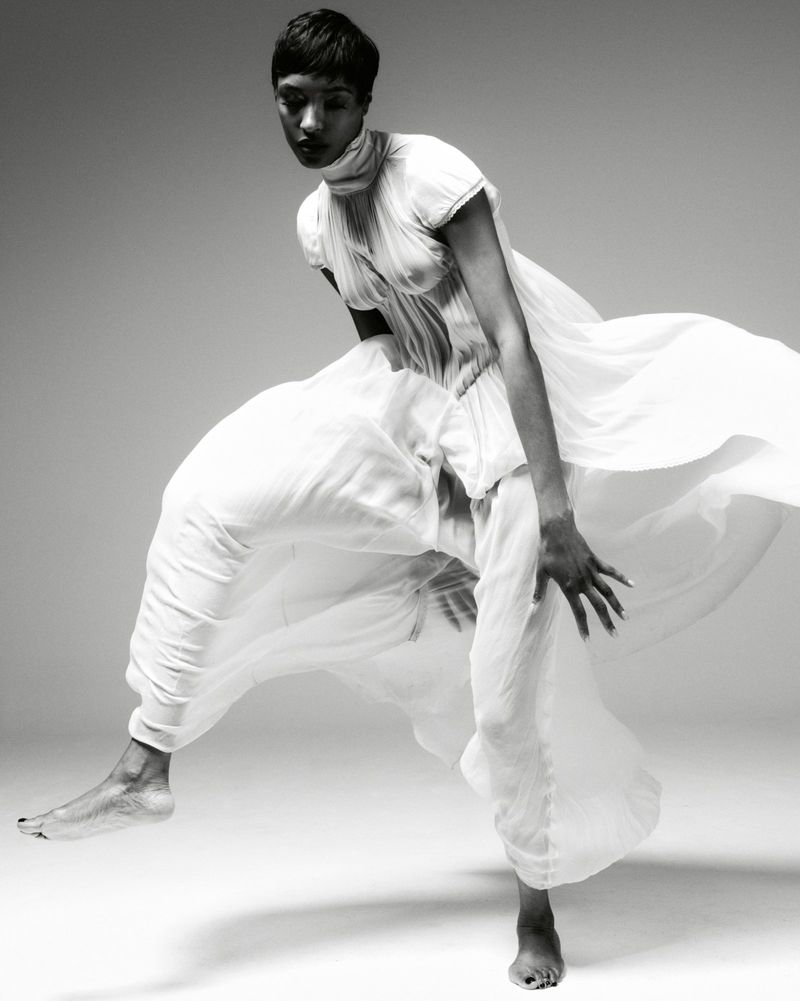 A black and white fashion shot of a woman in motion, wearing flowing white clothing, captured in a photo studio with soft, diffused lighting.