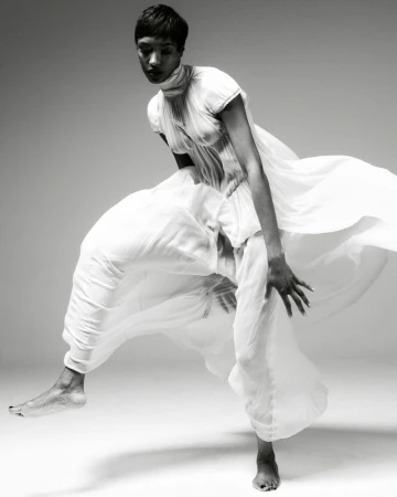 A black and white fashion shot of a woman in motion, wearing flowing white clothing, captured in a photo studio with soft, diffused lighting.