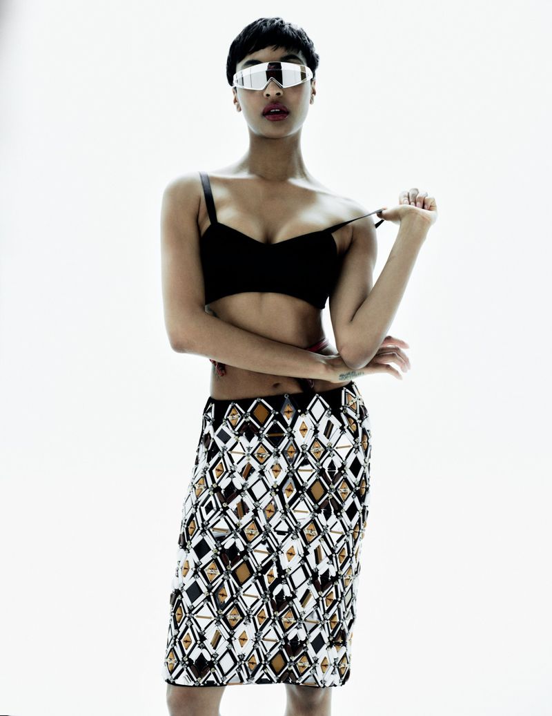 Portrait of a woman in a black bra, patterned skirt, and reflective sunglasses, standing against a bright white background.