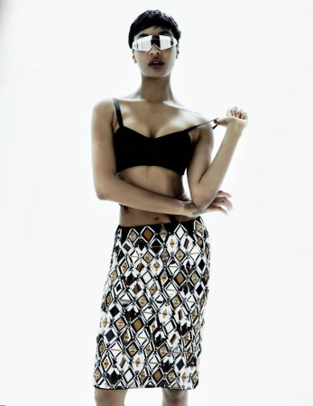 Portrait of a woman in a black bra, patterned skirt, and reflective sunglasses, standing against a bright white background.