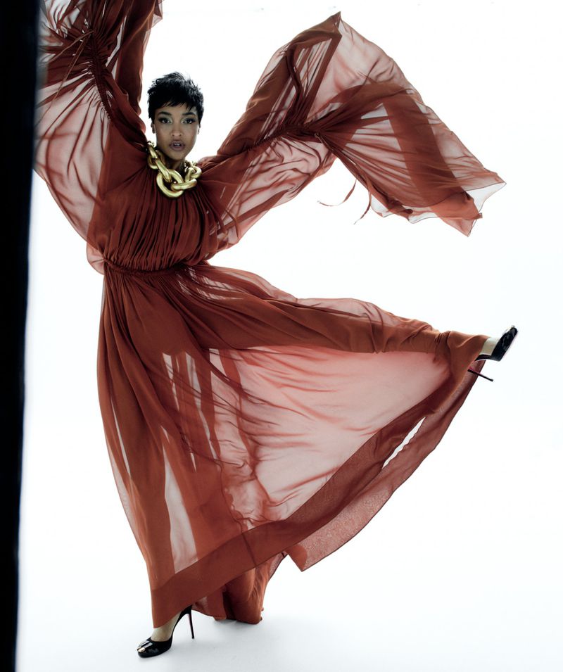 Elegant woman in motion wearing a flowing terracotta dress and gold necklace against a white background in a fashion style photo.