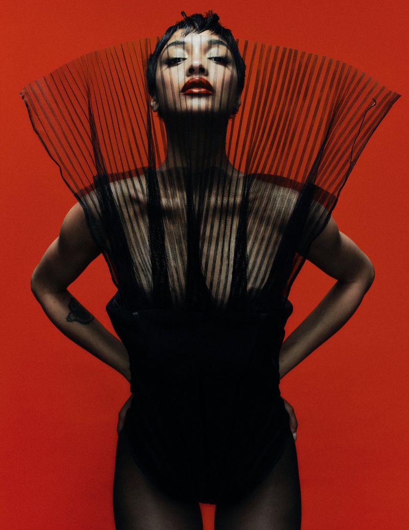 Fashion portrait of a woman with dark hair and red lips in a black outfit with a sheer, pleated collar, posed against a red backdrop.