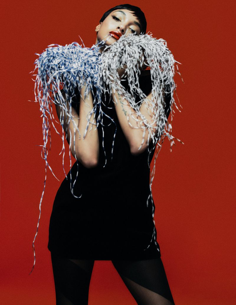 A woman in a black dress poses in front of a red backdrop, adorned with an intricate feather boa and striking red lipstick.