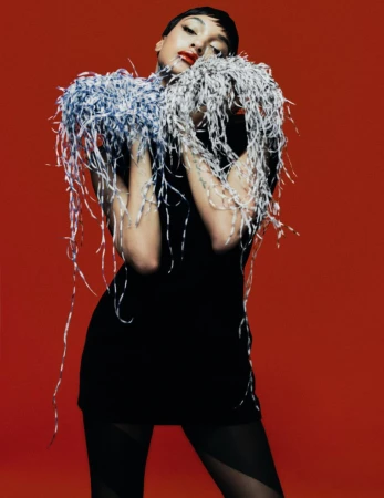A woman in a black dress poses in front of a red backdrop, adorned with an intricate feather boa and striking red lipstick.