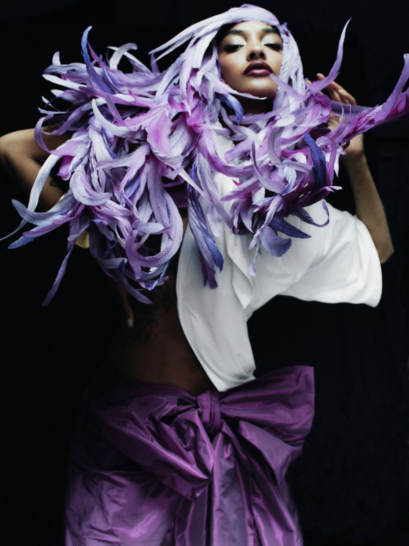 Fashion shot of a woman wearing feathers and a purple gown with a bow, posing against a black background with bright studio lighting.