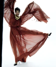 Model Jourdan Dunn in a flowing brown chiffon gown and chunky gold necklace dancing against a plain white studio backdrop.